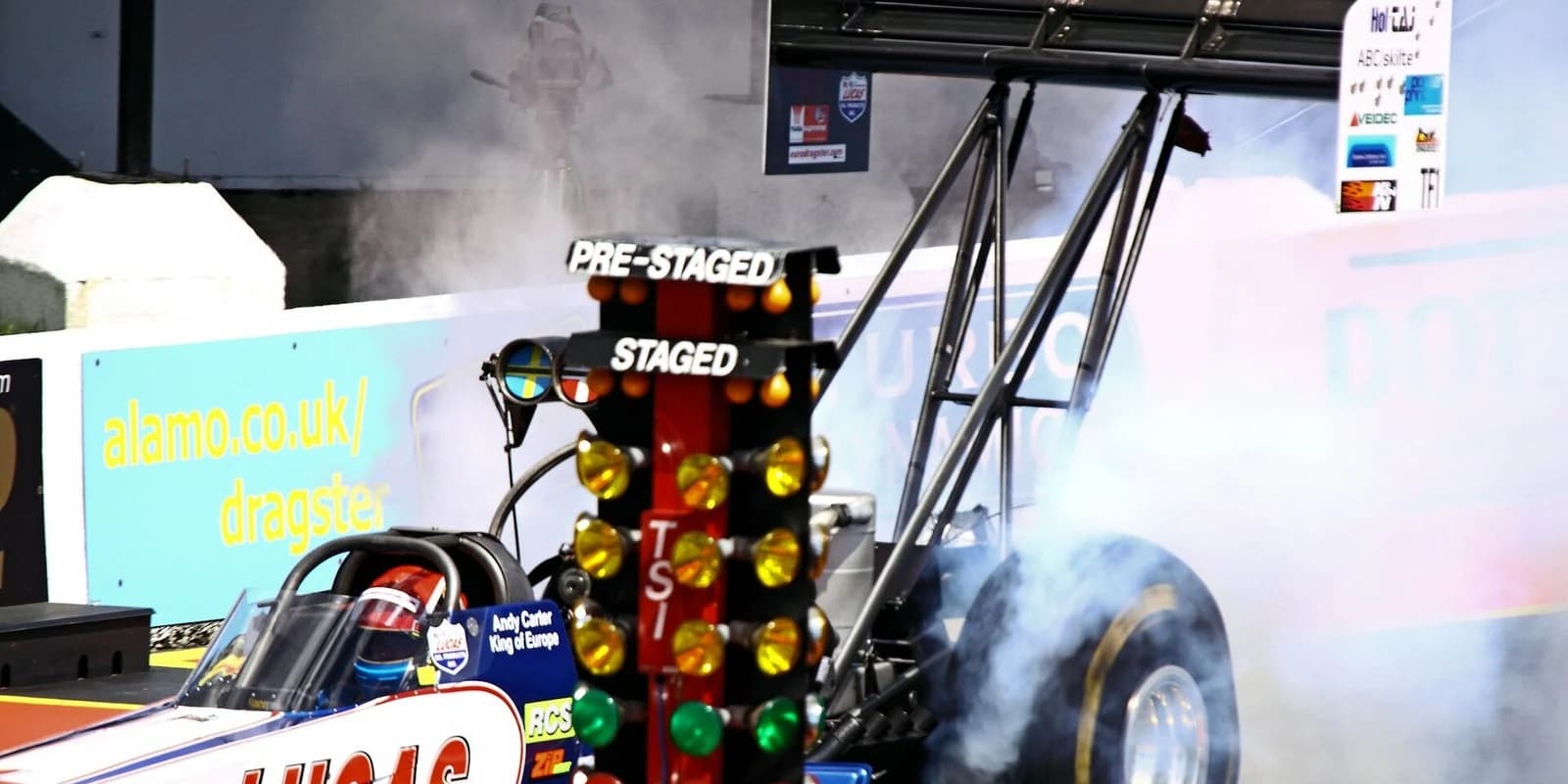 A Top Fuel dragster deep in a burnout at the startline Christmas tree, tyre smoke rolling off the rear slick, at Santa Pod Raceway