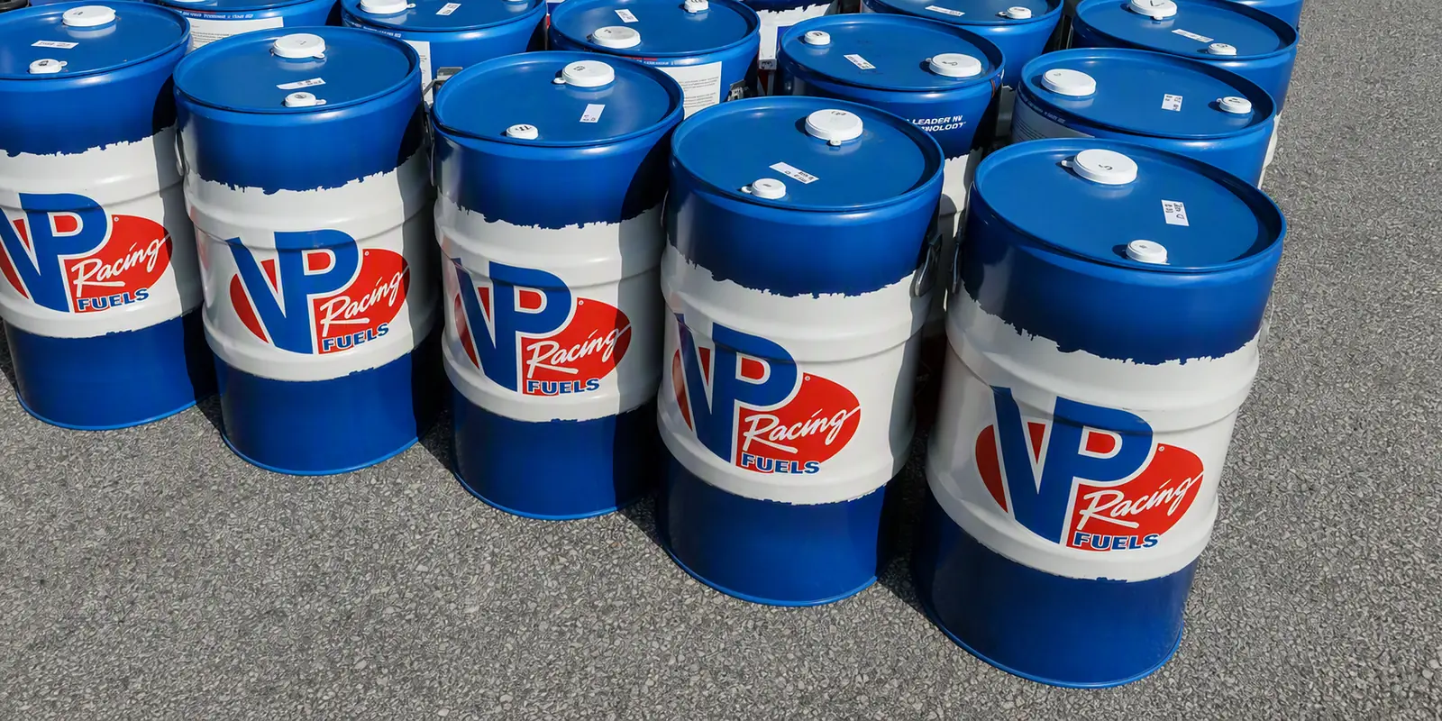 A dozen sealed blue and white VP Racing Fuels drums standing together on tarmac, each with a small hand-marked label on the lid