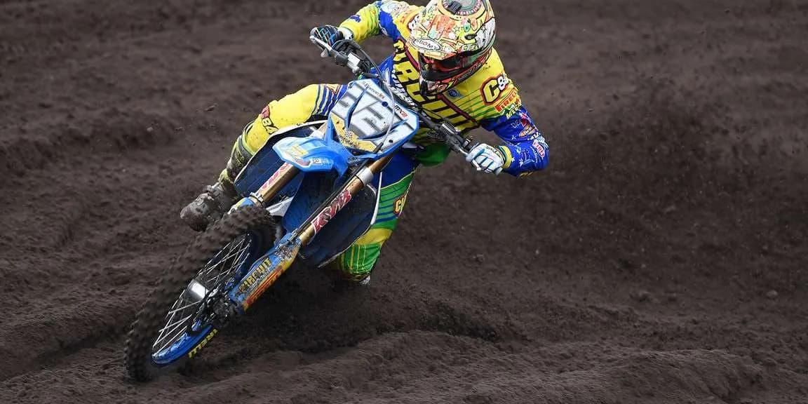 A rider cornering hard through deep sand on a blue-plasticked machine in yellow and green kit, the front number plate carrying an MX2 class marking