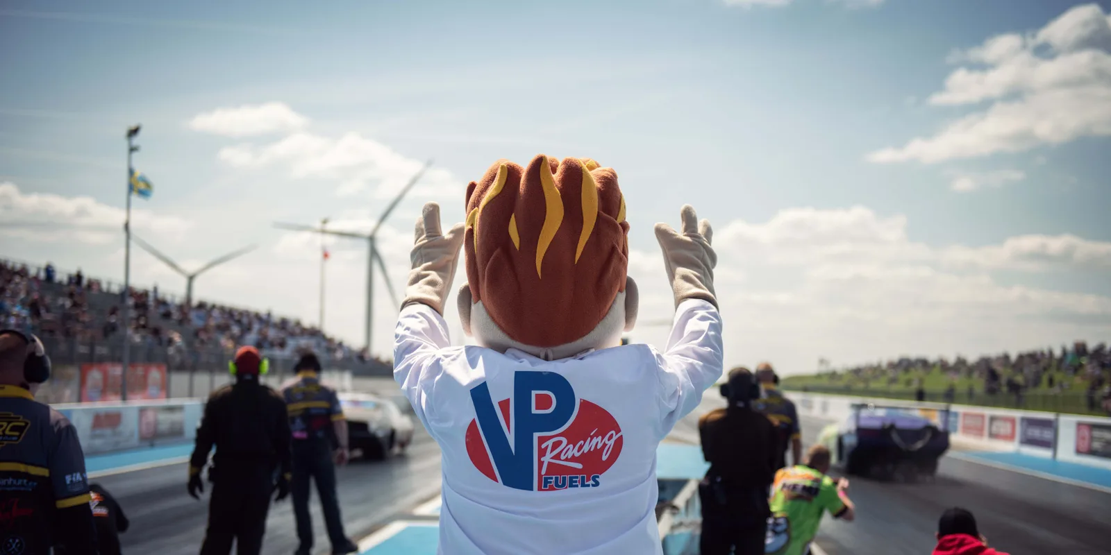 VP Racing's mascot, arms raised, facing a packed grandstand at a European drag strip, wind turbines and national flags behind